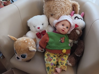 Na fotelu są umieszczone 3 pluszowe misie i 2 pluszowe pieski, na pluszowych maskotkach jest oparta lalka imitująca niemowlę w zielonym sweterku.