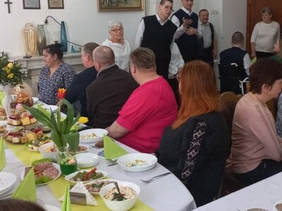 Czworo mieszkańców wykonuje część artystyczną, a pracownicy i pozostali mieszkańcy DPS siedzą za stołem i słuchają występ.