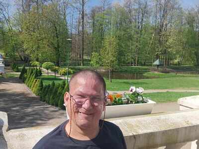 Mężczyzna usmiecha się prz bratkach na balkonie, a za balkonem widać zielony park.