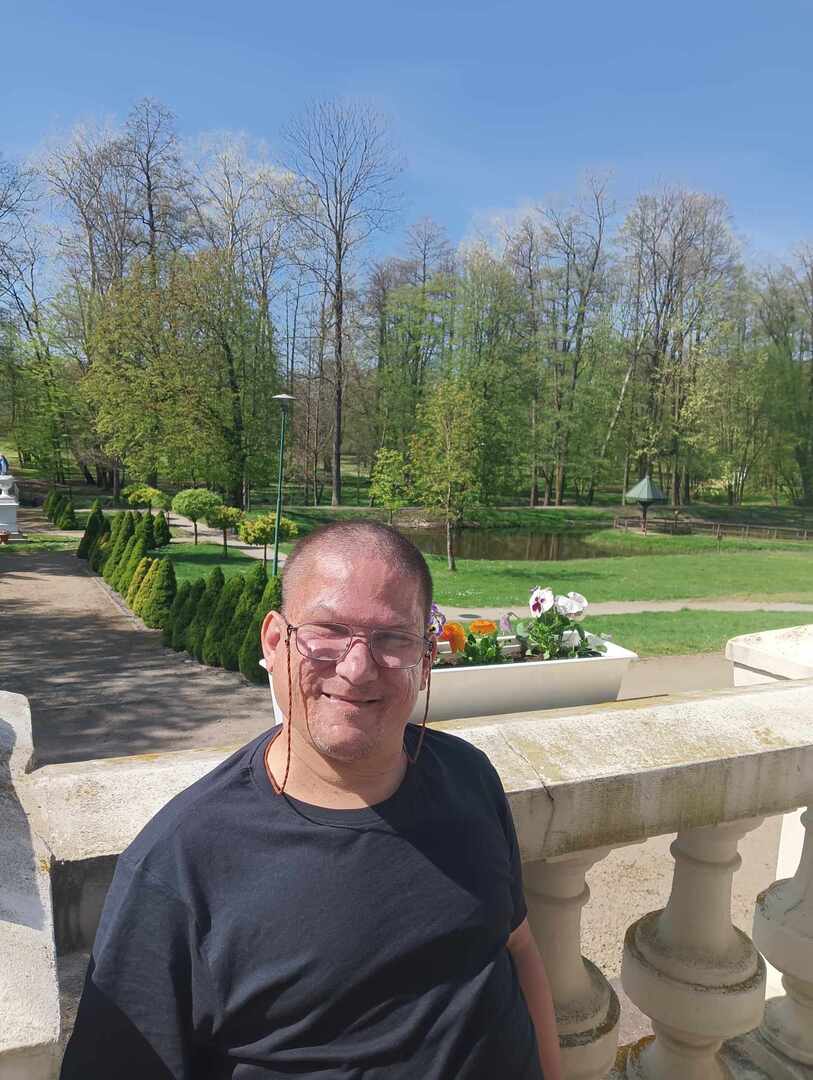 Mężczyzna usmiecha się prz bratkach na balkonie, a za balkonem widać zielony park.