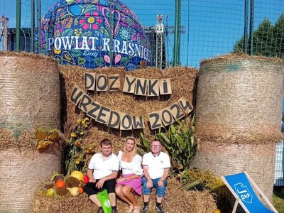 Dwóch mężczyzn i kobieta siedzą na belach siana wśród ozdób dożynkowych. Za plecami na belach siana jest napis: "Dożynki Urzędów 2024. Za belami siana widać balon reklamowy z napisem Powiat kraśnicki. 
