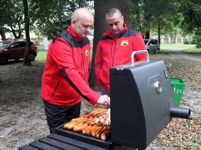 Dwóch panów z KGW Gościeradów pieką kiełbaski i szaszłyki na grilu.