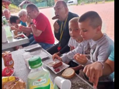 Uczestnicy biwaku siędzą za stołem i konsumeją poczęstunek z grilla