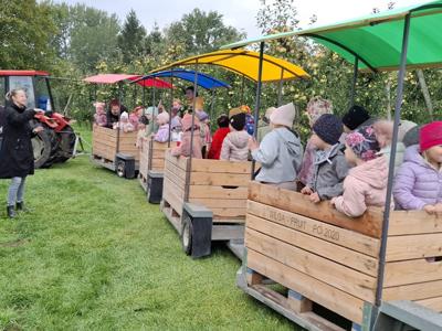 Dzieci w kolorowych kurtkach jadą drewnianą przyczepą z daszkiem przez sad.