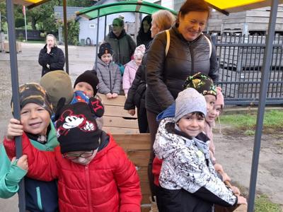 Dzieci w kolorowych kurtkach jadą drewnianą przyczepą z daszkiem przez sad.