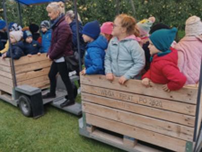 Dzieci w kolorowych kurtkach jadą drewnianą przyczepą z daszkiem przez sad.