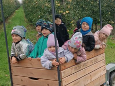 Dzieci w kolorowych kurtkach jadą drewnianą przyczepą z daszkiem przez sad.