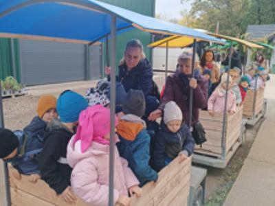 Dzieci w kolorowych kurtkach jadą drewnianą przyczepą z daszkiem przez sad.