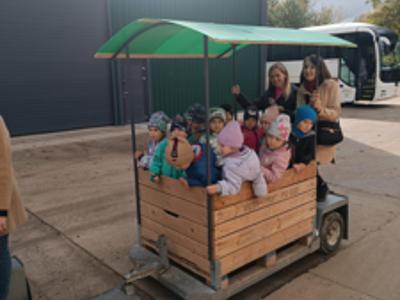 Dzieci w kolorowych kurtkach jadą drewnianą przyczepą z daszkiem przez sad.