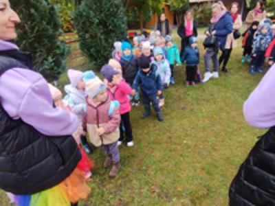 Grupa dzieci w jesiennych ubraniach, ustawiona w kolejce na świeżym powietrzu, otoczona drzewami i opiekunami.