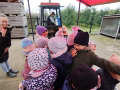 Dzieci w kolorowych kurtkach podczas przejażdżki drewnianą przyczepą przez sad.