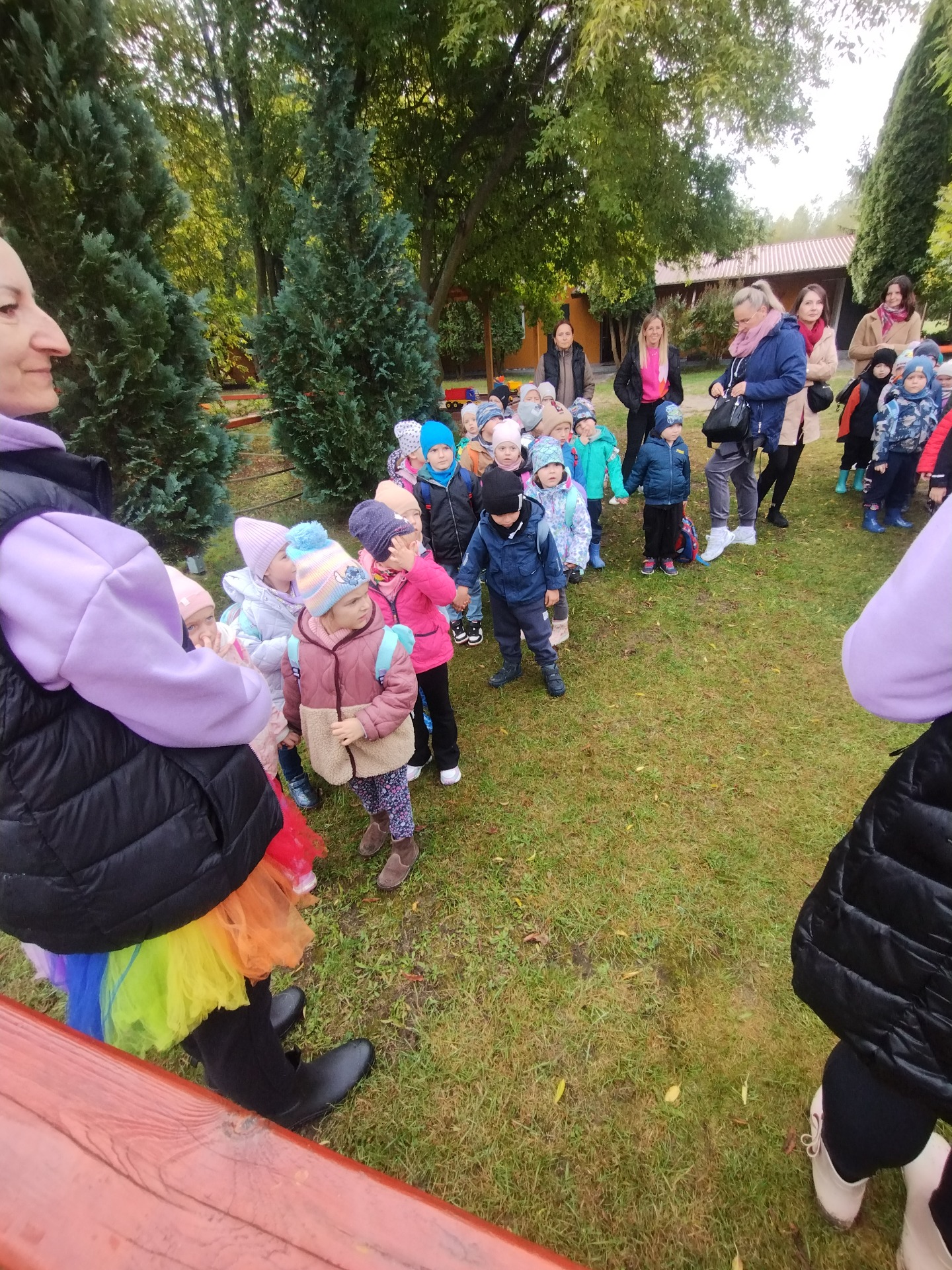 Grupa dzieci w jesiennych ubraniach, ustawiona w kolejce na świeżym powietrzu, otoczona drzewami i opiekunami.