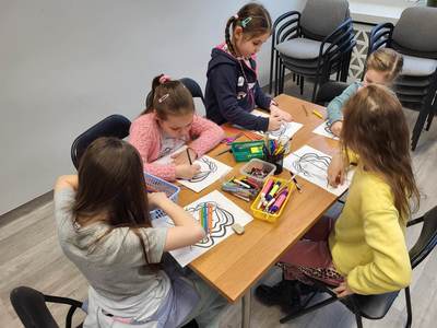 Zdjęcie przedstawia dzieci uczęszczające na warsztaty. Dzieci siedzą przy stole i wykonują prace plastyczne.