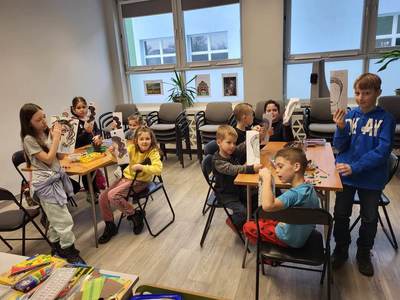 Zdjęcie przedstawia dzieci uczęszczające na warsztaty. Dzieci stoją, siedzą przy stole i wykonują prace plastyczne.