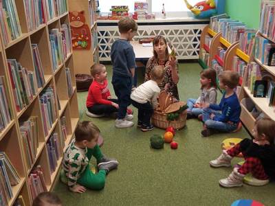 Na zdjęciu znajdują się uczestnicy warsztatów, dzieci siedzą na poduszkach. W tle księgozbiór biblioteki.