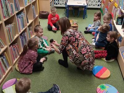 Na zdjęciu znajdują się uczestnicy warsztatów, dzieci siedzą na poduszkach. W tle księgozbiór biblioteki.