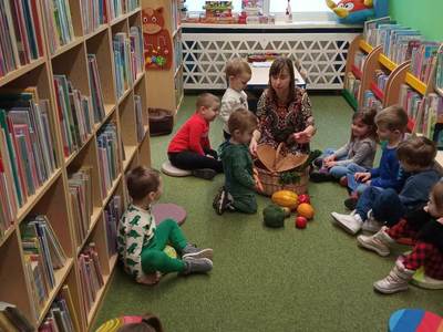 Na zdjęciu znajdują się uczestnicy warsztatów, dzieci siedzą na poduszkach. W tle księgozbiór biblioteki.