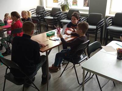 Na zdjęciu znajdują się uczestnicy warsztatów. Dzieci siedzą przy stole i wykonują prace plastyczne.