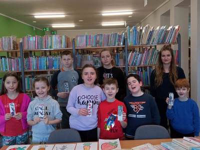 Na zdjęciu znajdują się uczestnicy zajęć trzymający w dłoni prace plastyczne. W tle księgozbiór biblioteki.