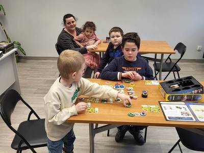 Na zdjęciu znajdują się uczestnicy bibliotecznych spotkań. Uczestnicy siedzą przy stole i grają w gry planszowe.