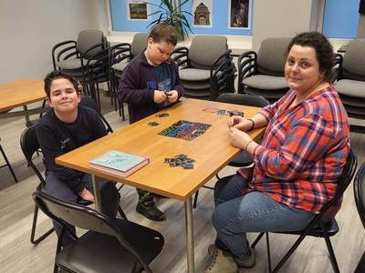 Na zdjęciu znajdują się uczestnicy bibliotecznych spotkań. Uczestnicy siedzą przy stole i grają w gry planszowe.