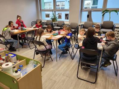 Na zdjęciu znajdują się uczestnicy warsztatów. Dzieci siedzą przy stołach i wykonują prace plastyczne.