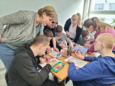 Na zdjęciu znajdują się uczestnicy warsztatów. Młodzież siedzi przy stole i wykonuje prace plastyczne.