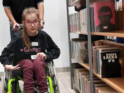 Na zdjęciu znajdują się uczestnicy warsztatów. Dziewczynka siedzi na wózku inwalidzkim pomiędzy regałami z książkami.