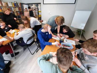 Na zdjęciu znajdują się uczestnicy warsztatów. Młodzież siedzi przy stole i wykonuje prace plastyczne.
