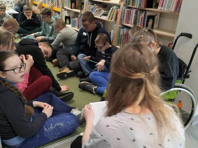 Na zdjęciu znajdują się uczestnicy warsztatów. Młodzież siedzi na poduszkach pomiędzy regałami.