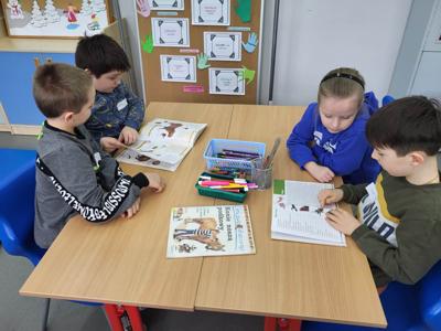 Na zdjęciu znajdują się uczestnicy zajęć. Dzieci siedzą na krzesłach w przy stołach i czytają książki.