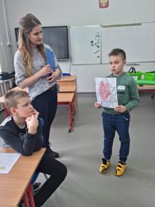 Na zdjęciu znajdują się uczestnicy zajęć. Dziecko pokazuje wykonaną prace plastyczną.