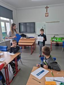 Na zdjęciu znajdują się uczestnicy zajęć. Dziecko pokazuje wykonaną prace plastyczną.