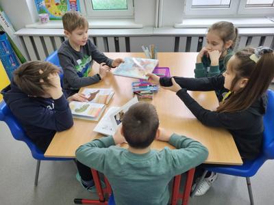Na zdjęciu znajdują się uczestnicy zajęć. Dzieci siedzą na krzesłach w przy stołach i czytają książki.