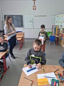 Na zdjęciu znajdują się uczestnicy zajęć. Dziecko pokazuje wykonaną prace plastyczną.