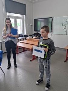 Na zdjęciu znajdują się uczestnicy zajęć. Dziecko pokazuje wykonaną prace plastyczną.