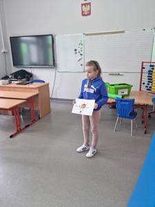 Na zdjęciu znajdują się uczestnicy zajęć. Dziecko pokazuje wykonaną prace plastyczną.