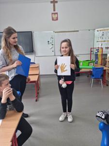 Na zdjęciu znajdują się uczestnicy zajęć. Dziecko pokazuje wykonaną prace plastyczną.