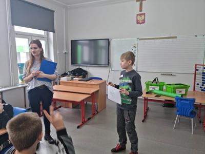 Na zdjęciu znajdują się uczestnicy zajęć. Dziecko pokazuje wykonaną prace plastyczną.