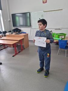 Na zdjęciu znajdują się uczestnicy zajęć. Dziecko pokazuje wykonaną prace plastyczną.