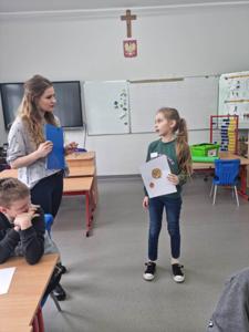 Na zdjęciu znajdują się uczestnicy zajęć. Dziecko pokazuje wykonaną prace plastyczną.