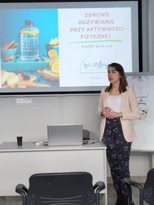 Na zdjęciu znajduje się prowadząca warsztat. W tle wyświetlana prezentacja multimedialna. Zdjęcie wykonano w w bibliotece w sali wielofunkcyjnej.