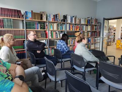 Na zdjęciu znajdują się uczestnicy spotkania. Uczestnicy siedzą na krzesłach w tle księgozbiór biblioteki.