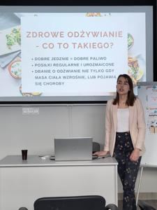 Na zdjęciu znajduje się prowadząca warsztat. W tle wyświetlana prezentacja multimedialna. Zdjęcie wykonano w w bibliotece w sali wielofunkcyjnej.