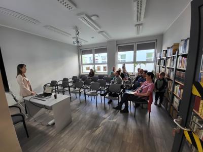 Na zdjęciu znajdują się uczestnicy spotkania. Uczestnicy siedzą na krzesłach w tle księgozbiór biblioteki.