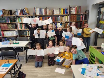 Na zdjęciu znajdują się uczestnicy zajęć. Dzieci trzymają w rękach prace plastyczne. Zdjęcie wykonano w sali wielofunkcyjnej biblioteki.