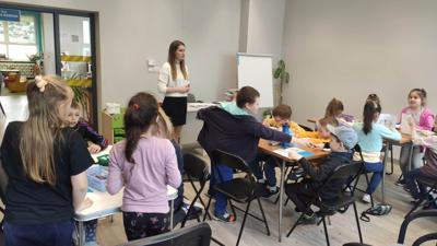 Na zdjęciu znajdują się uczestnicy zajęć. Dzieci wykonują prace plastyczne siedząc przy stole. Zdjęcie wykonano w sali wielofunkcyjnej biblioteki.