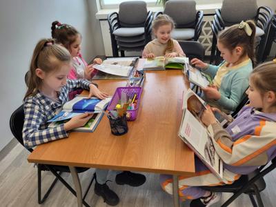 Na zdjęciu znajdują się uczestnicy zajęć. Dzieci siedzą przy stole i czytają książki. Zdjęcie wykonano w sali wielofunkcyjnej biblioteki.