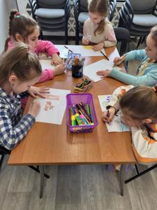 Na zdjęciu znajdują się uczestnicy zajęć. Dzieci wykonują prace plastyczne siedząc przy stole. Zdjęcie wykonano w sali wielofunkcyjnej biblioteki.