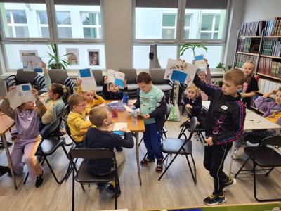 Na zdjęciu znajdują się uczestnicy zajęć. Dzieci siedzą przy stole i pokazują prace plastyczne. Zdjęcie wykonano w sali wielofunkcyjnej biblioteki.